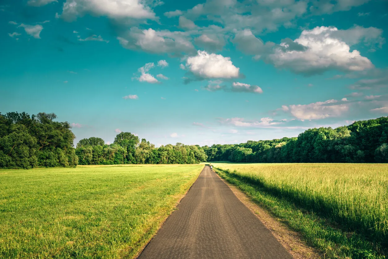 A paved path running between open fields beneath a blue, cloud-filled sky.